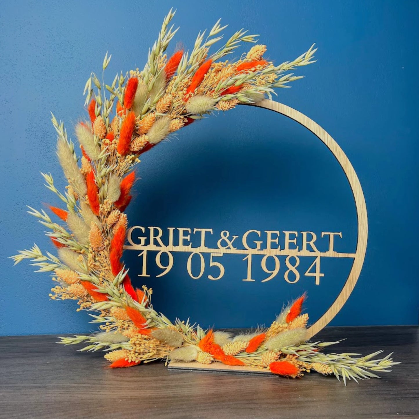 Couronne en bois ornée de fleurs séchées, personnalisée avec les prénoms "Griet & Geert" et la date "19 05 1984". La couronne est partiellement décorée de fleurs séchées orange et beige et de graminées, et est posée sur un socle en bois contre un fond bleu.
