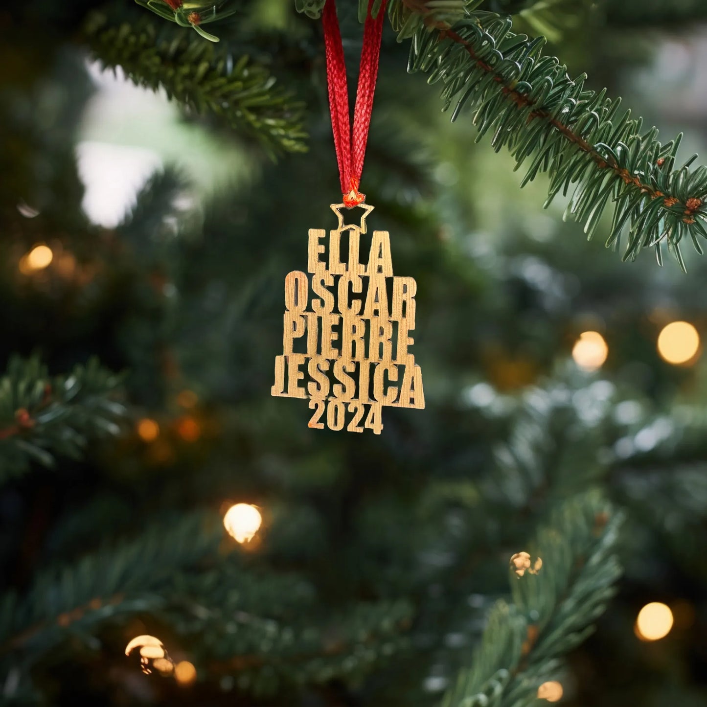 Une décoration en bois avec les prénoms "Ella", "Oscar", "Pierre", "Jessica" et l'année "2024", suspendue à un sapin de Noël avec un ruban rouge. La scène est éclairée par des lumières de Noël, créant une ambiance chaleureuse et festive dans un cadre naturel de branches de sapin.