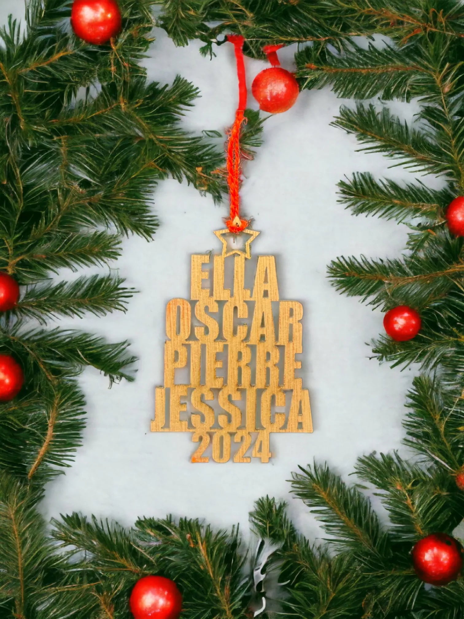 Décoration en bois suspendue, gravée avec les prénoms "Ella", "Oscar", "Pierre", "Jessica", et l'année "2024". L'ornement est entouré de branches de sapin avec des boules rouges, offrant une touche festive parfaite pour une décoration de Noël. Le contraste du bois clair et des ornements rouges ajoute un charme rustique et personnalisé à l'ensemble.